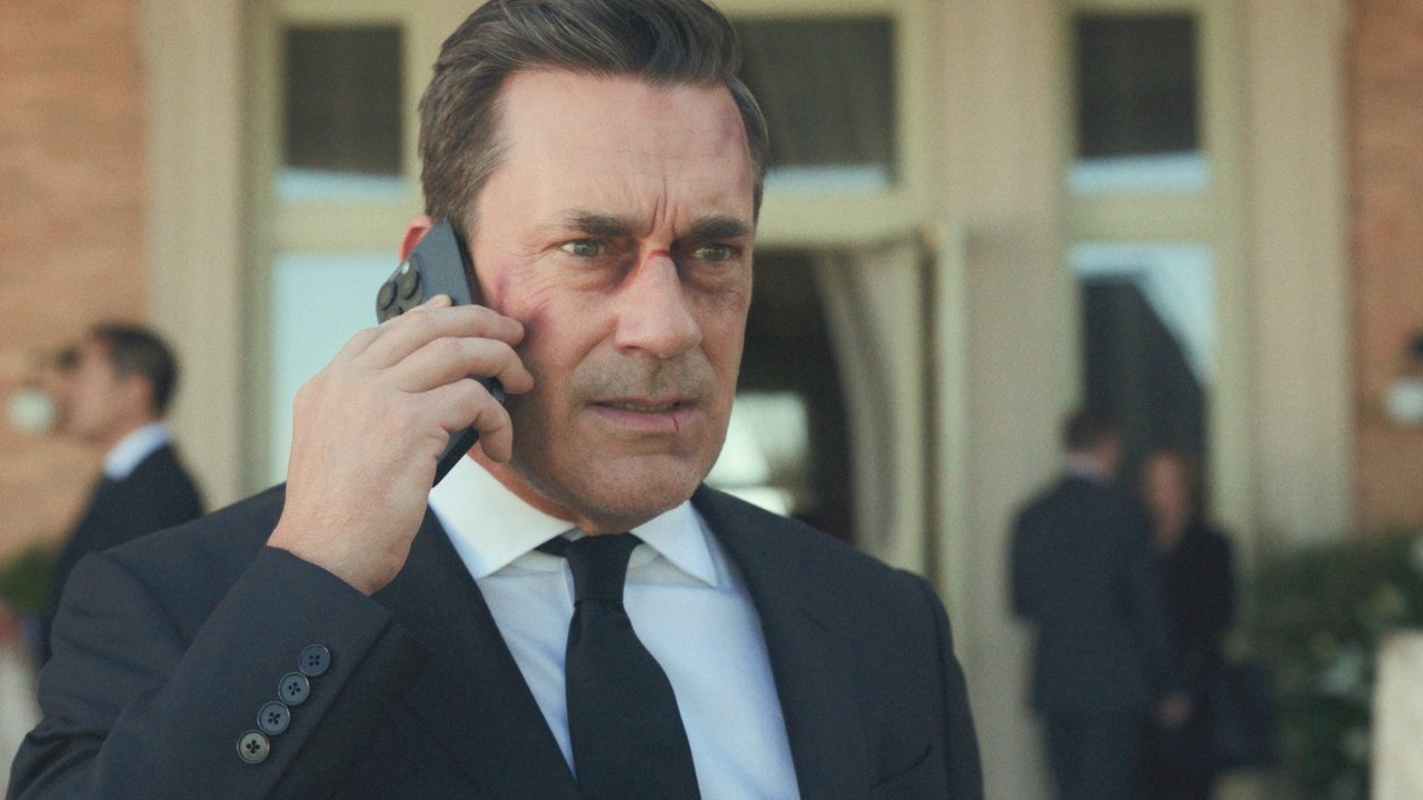Man in a suit with bruises on his face, talking on a cellphone, standing outside a building with people in the background.