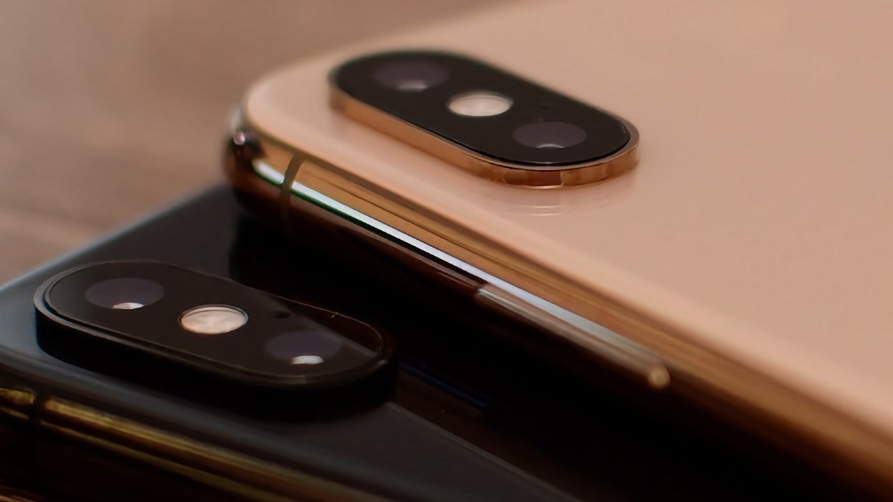 Close-up of two smartphones with dual rear cameras, one gold and one black, lying on a wooden surface.