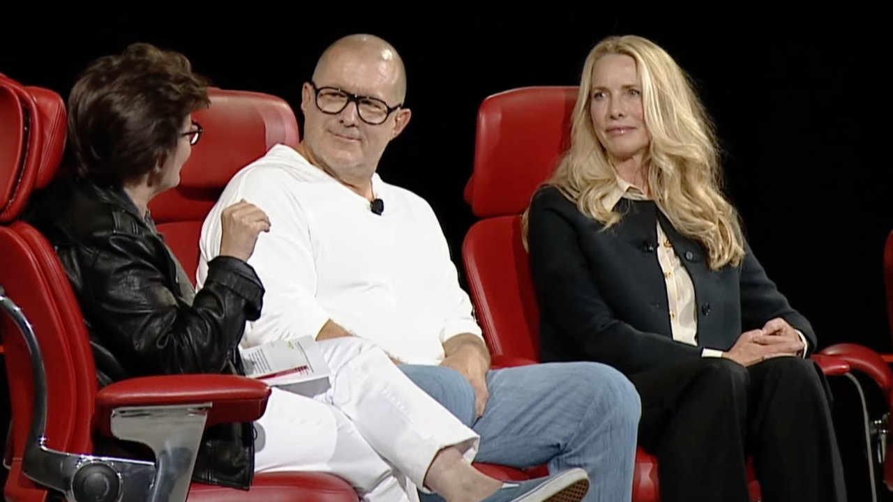 Three people sitting on red chairs, engaged in conversation. The person on the left wears glasses and a leather jacket, the middle person in white, and the right person in black.