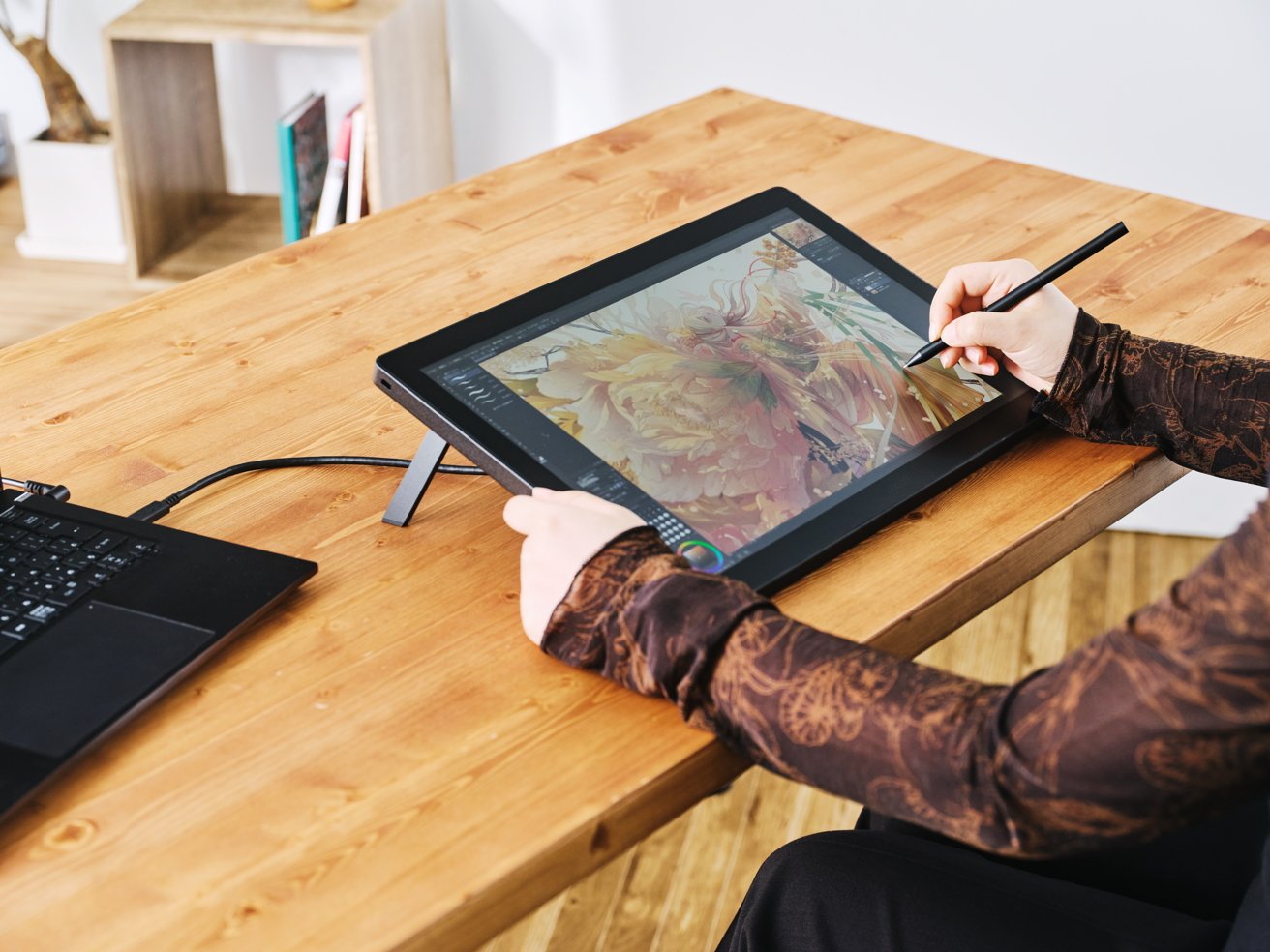 Person drawing digitally on a tablet with a stylus at a wooden desk, connected to a laptop. Shelves and books visible in the background.