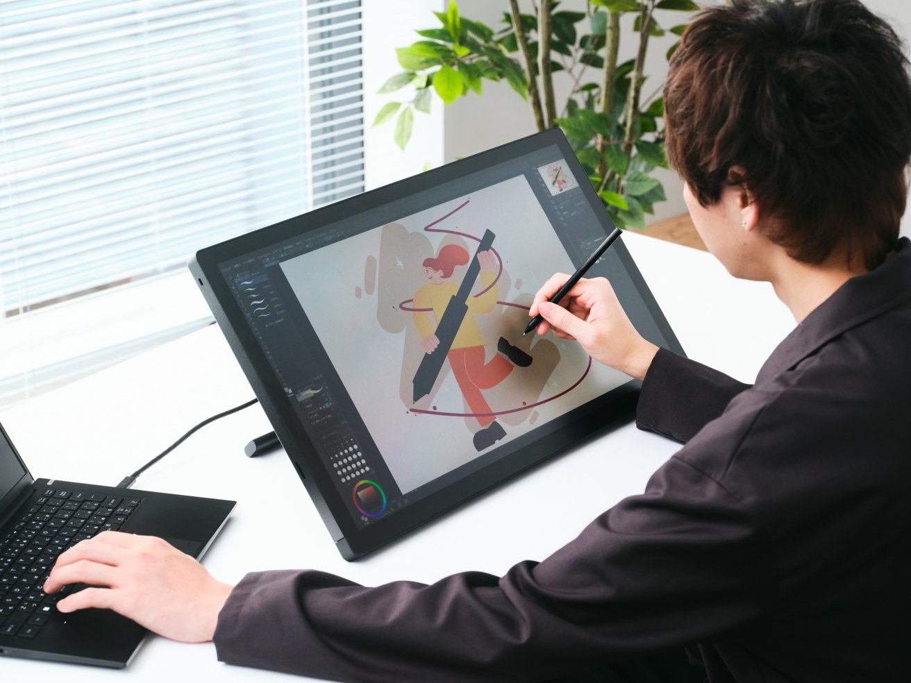 Person using a digital pen to draw colorful art on a large tablet, connected to a laptop, with a plant and window blinds in the background.
