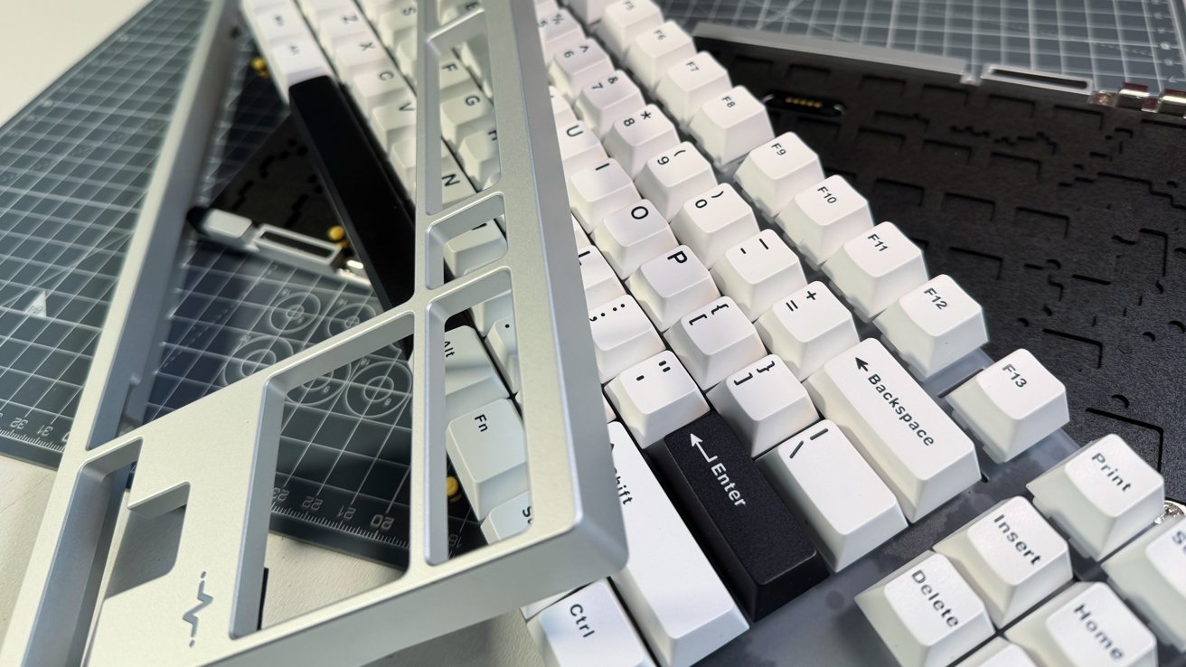 White keyboard keys with black labels on a grid-patterned mat, alongside a disassembled gray metallic frame.