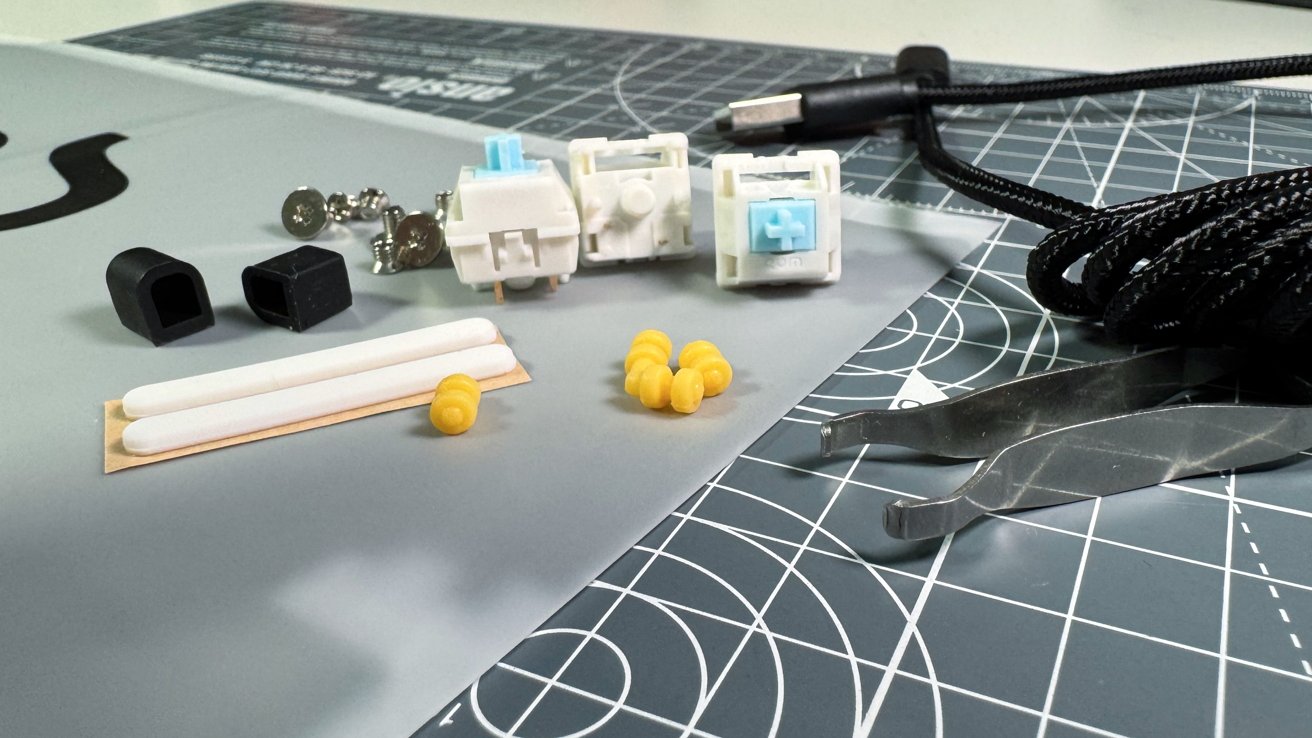 Mechanical keyboard switches, black keycap pullers, white swabs, yellow earplugs, silver tweezers, and a black braided cable on a grid cutting mat.