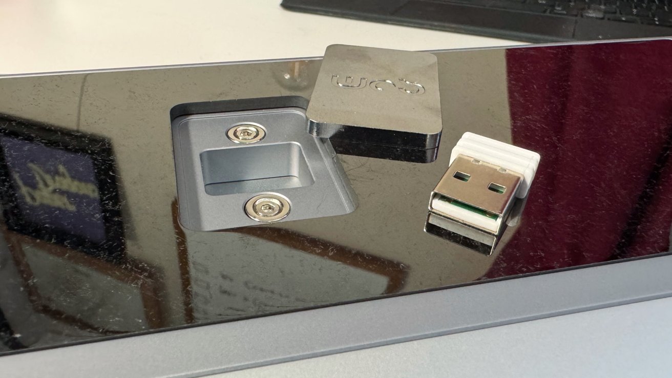 Close-up of a glossy black surface with a recessed slot, two screws, a USB dongle, and a metal square piece with engraved text.