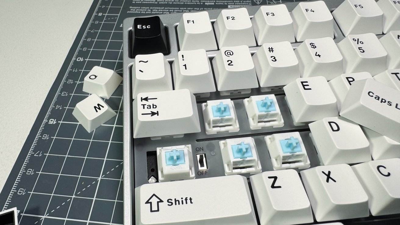White keyboard with blue switches, black Esc key, some keys removed, placed on a cutting mat with measurements.