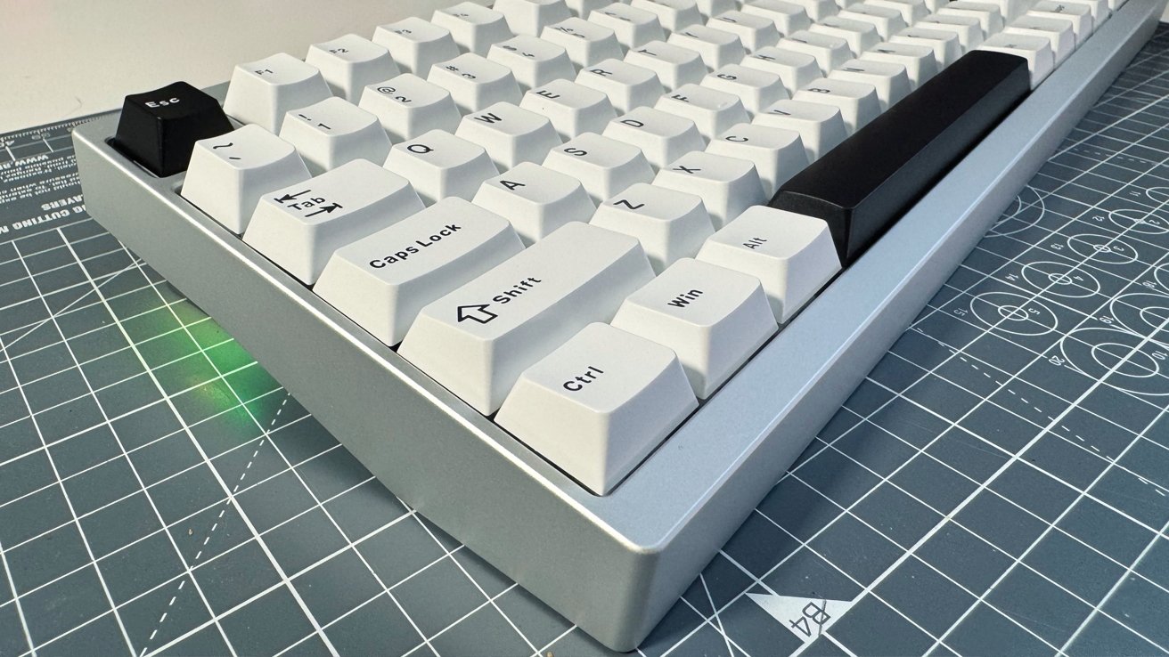 A mechanical keyboard with white keys and a black escape key on a blue grid-patterned cutting mat, lit by subtle green light.