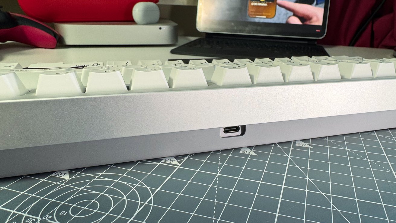 White keyboard on a gray grid-patterned mat, background showing red mouse, tablet, and a Mac Mini computer.