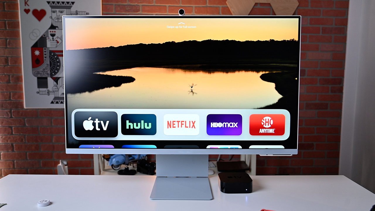 Samsung monitor displaying streaming apps like Apple TV, Hulu, and Netflix against a sunset lake scene, placed on a white desk with a brick wall background.