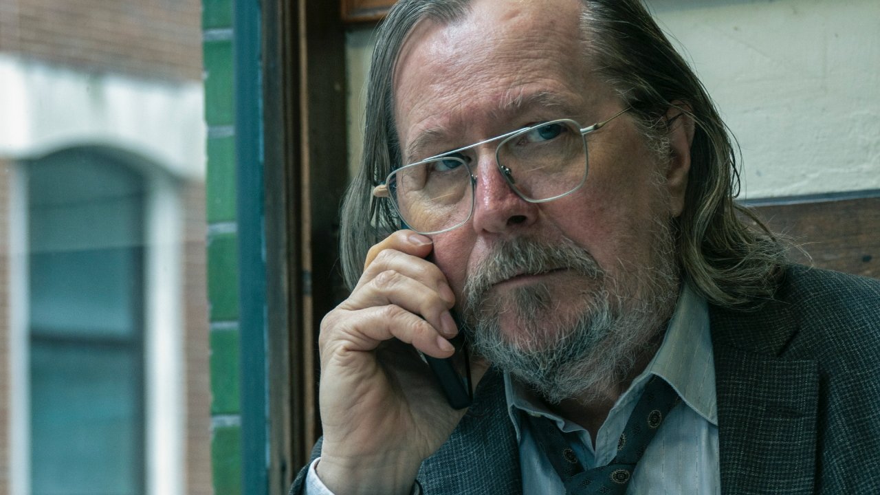 Older man with glasses and long hair, wearing a suit and tie, holding a phone to his ear while sitting indoors with a thoughtful expression.