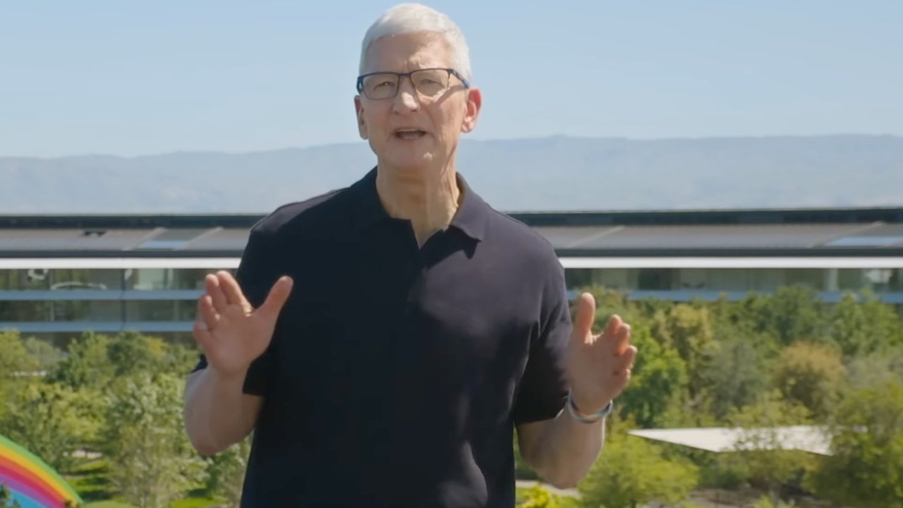 A person with short white hair and glasses speaks outdoors in front of a modern building and trees.