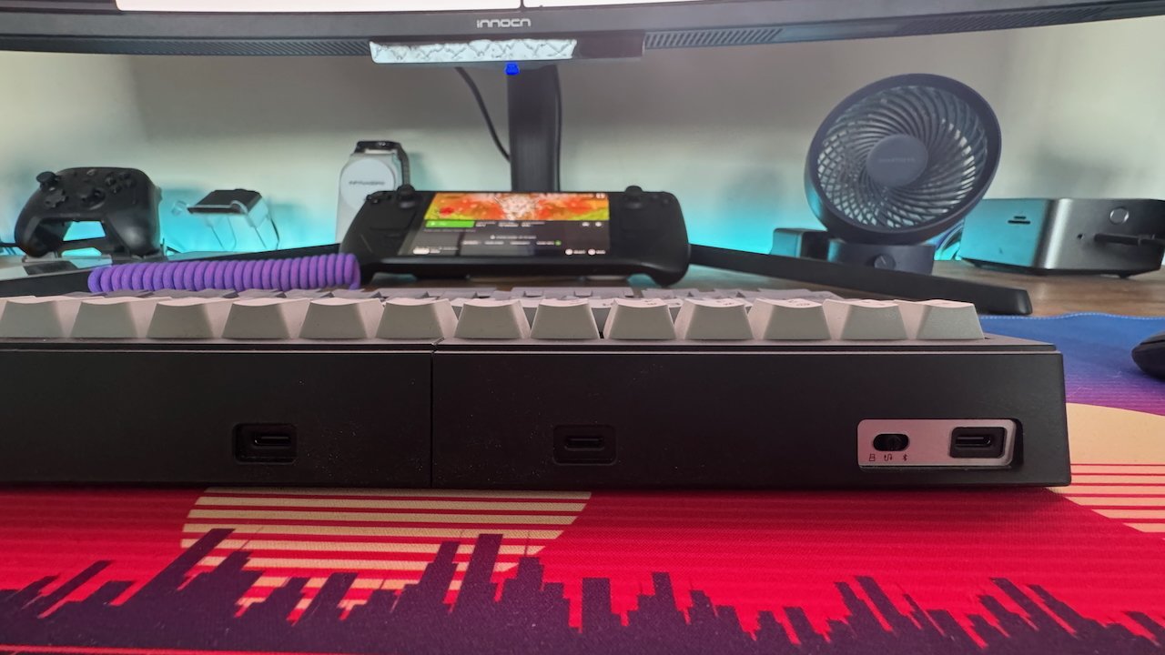 Computer setup with a keyboard, purple cable, and gaming controller on a colorful mat; a fan and monitor are in the background.