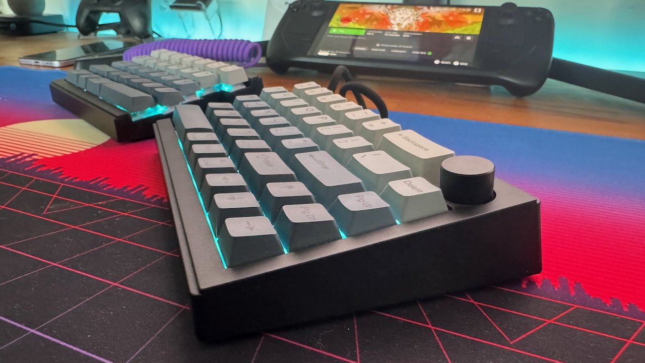 Mechanical keyboard with teal backlighting on a colorful mouse pad, purple cable, and handheld gaming console in the background.