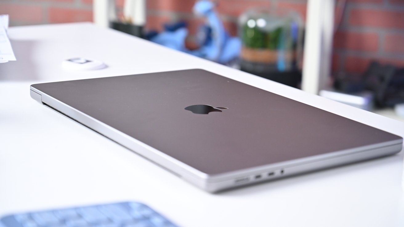 M3 MacBook Pro resting on a white desk, Apple logo visible, with a blurred background including a small white device and blue objects.
