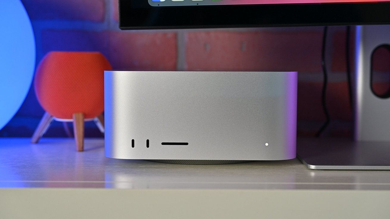 A silver Mac Studio on a table with a red smart speaker, in front of a brick wall and monitor screen.