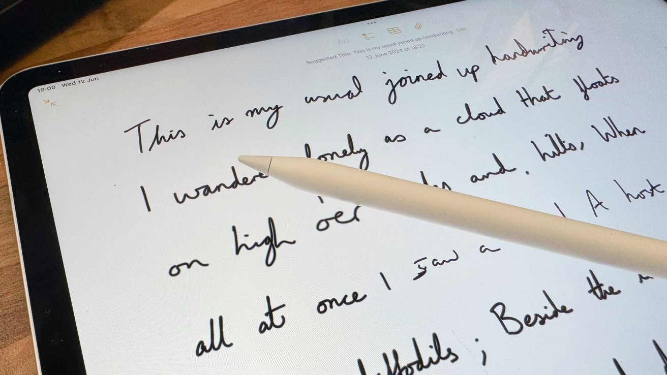 A tablet displaying cursive handwriting with a white stylus resting on its screen, set on a wooden surface.