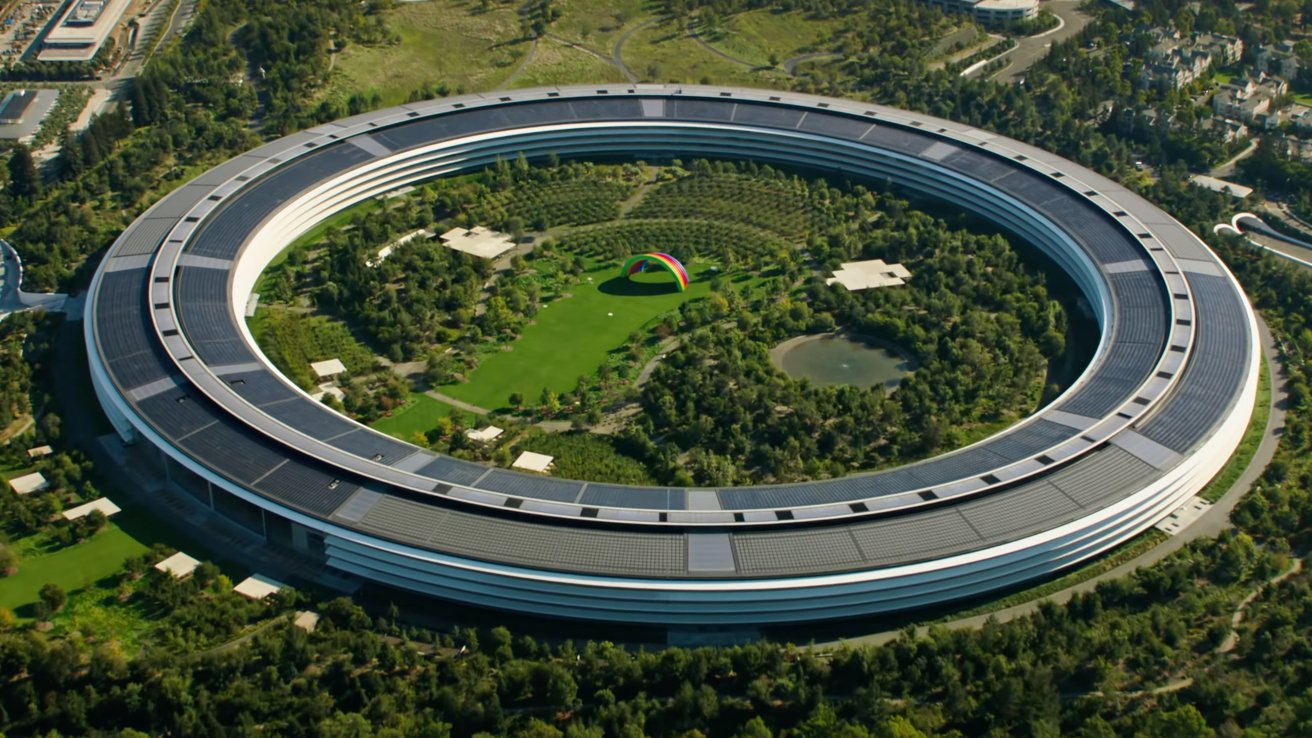 Aerial view of a circular building surrounded by trees, with a green courtyard in the center, featuring a colorful rainbow structure and a small pond.