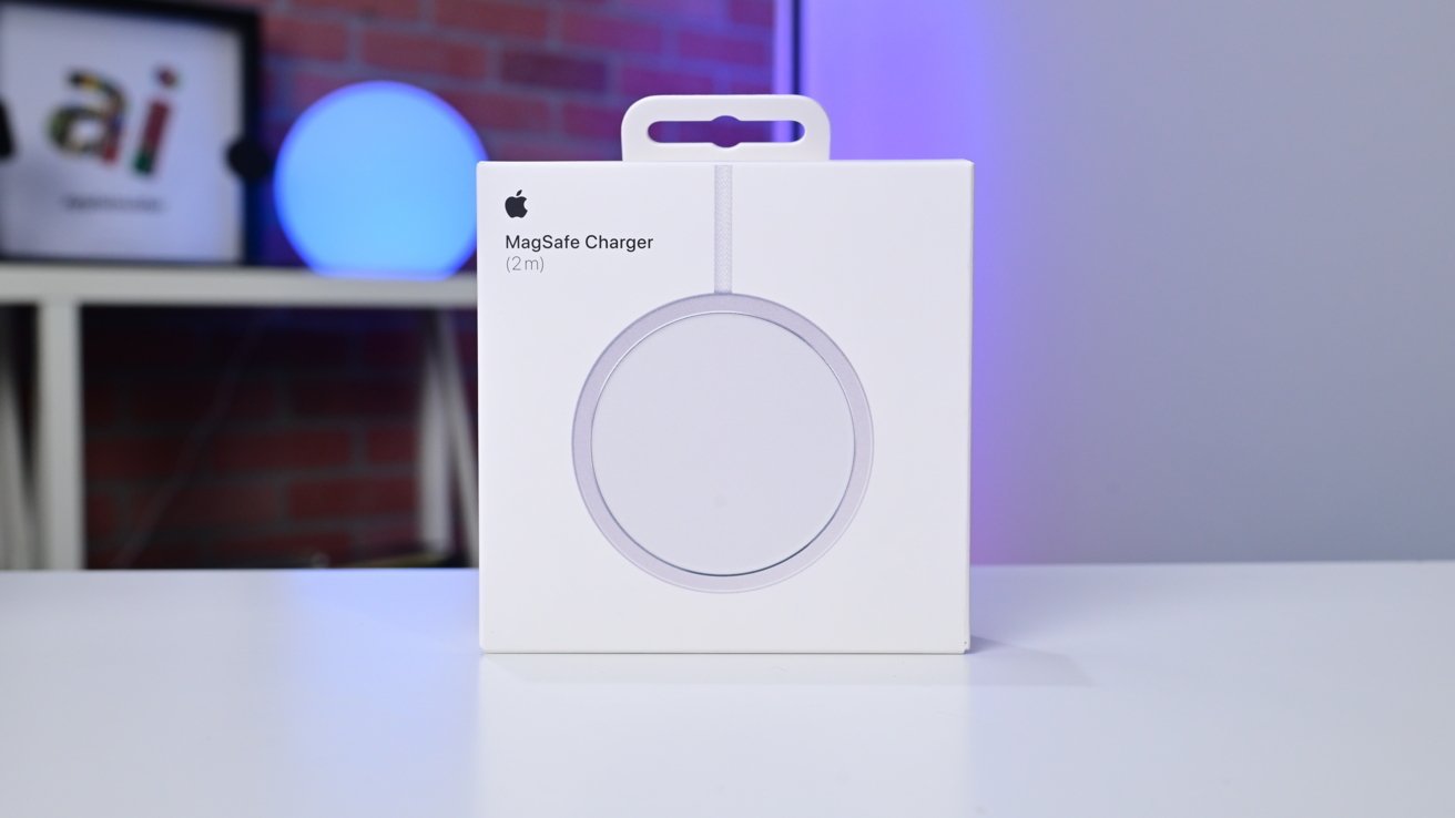 White MagSafe Charger box standing on a table, with shelves and a blue-lit sphere in the blurred background.