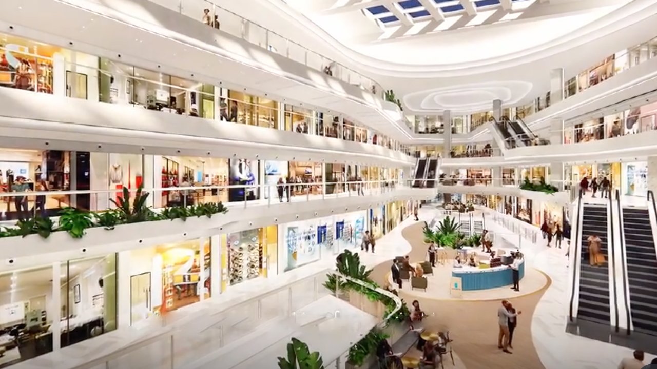 Spacious multi-level shopping mall with escalators, bright lighting, indoor plants, and people exploring various shops and cafes. Skylights illuminate the open, modern design.