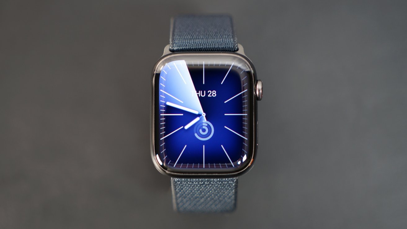 Smartwatch with a blue face displaying time and date, attached to a blue fabric strap against a dark background.