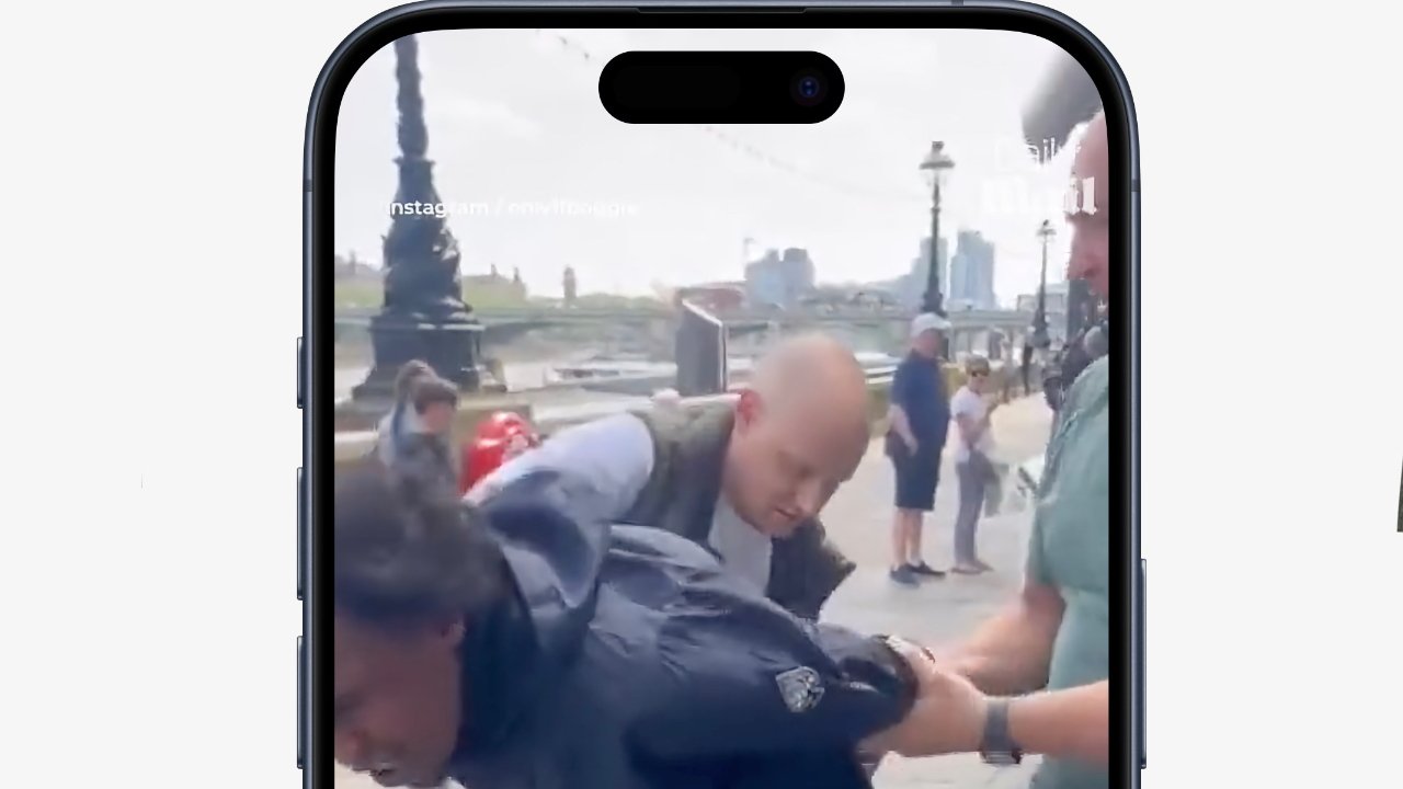 A person restrained by two others on a riverside walkway, with a bridge and people in the background. Displayed on a smartphone.