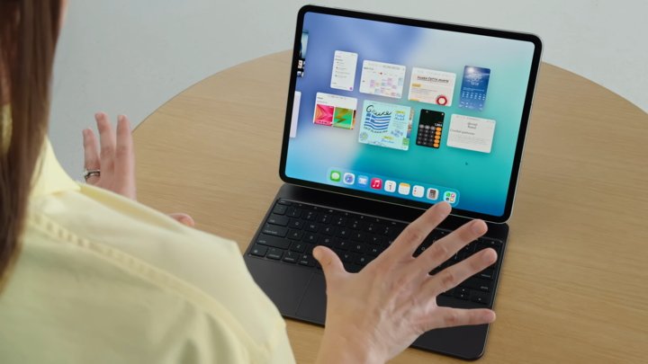 A person interacts with a tablet on a keyboard, displaying various app windows on-screen, seated at a wooden table.
