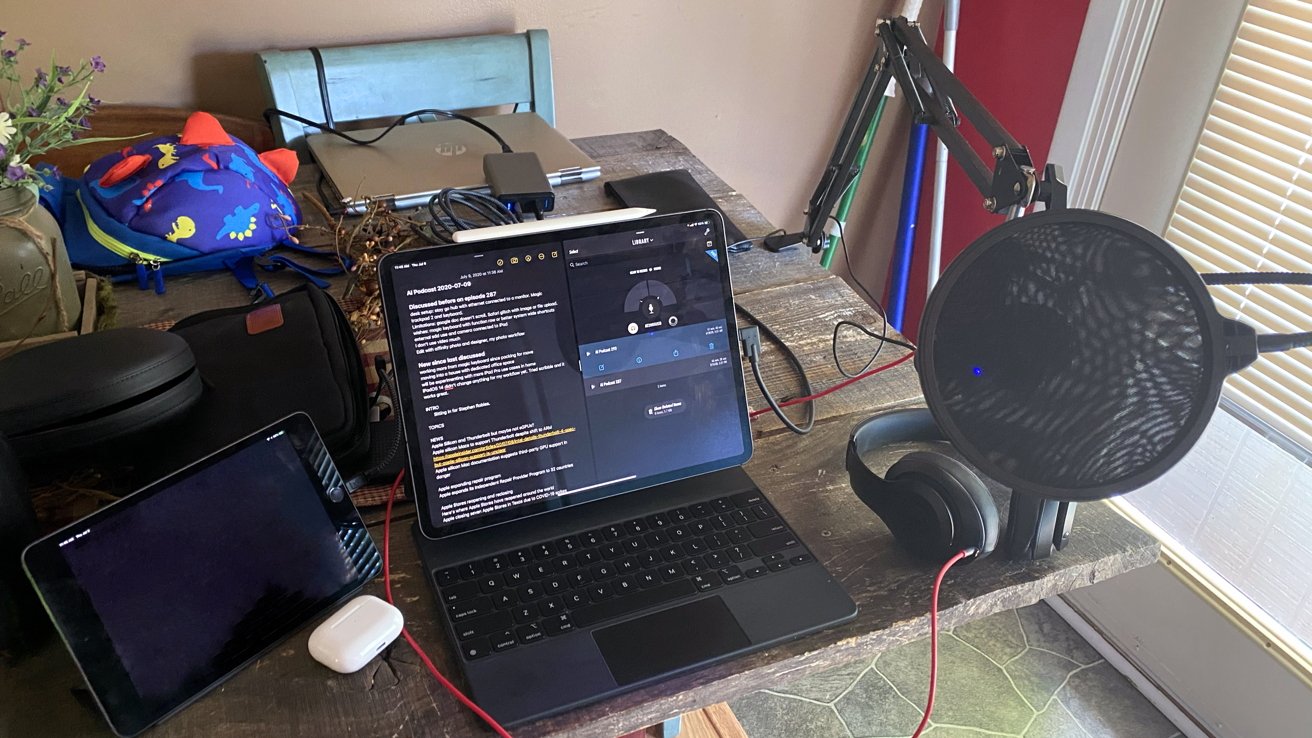 Home office setup with a tablet, laptop displaying text, headphones, microphone, and colorful dinosaur backpack on a wooden desk near a window.