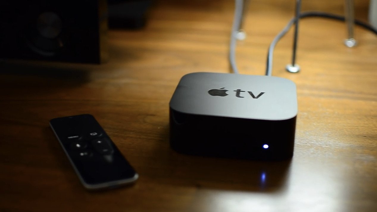 Apple TV device and remote on a wooden surface, with cables connected and a small light glowing on the device.