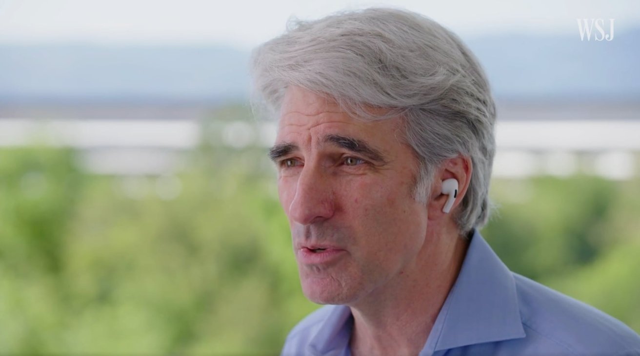 Man with gray hair wearing a blue shirt and white earbuds, speaking outdoors with trees and sky in the background.
