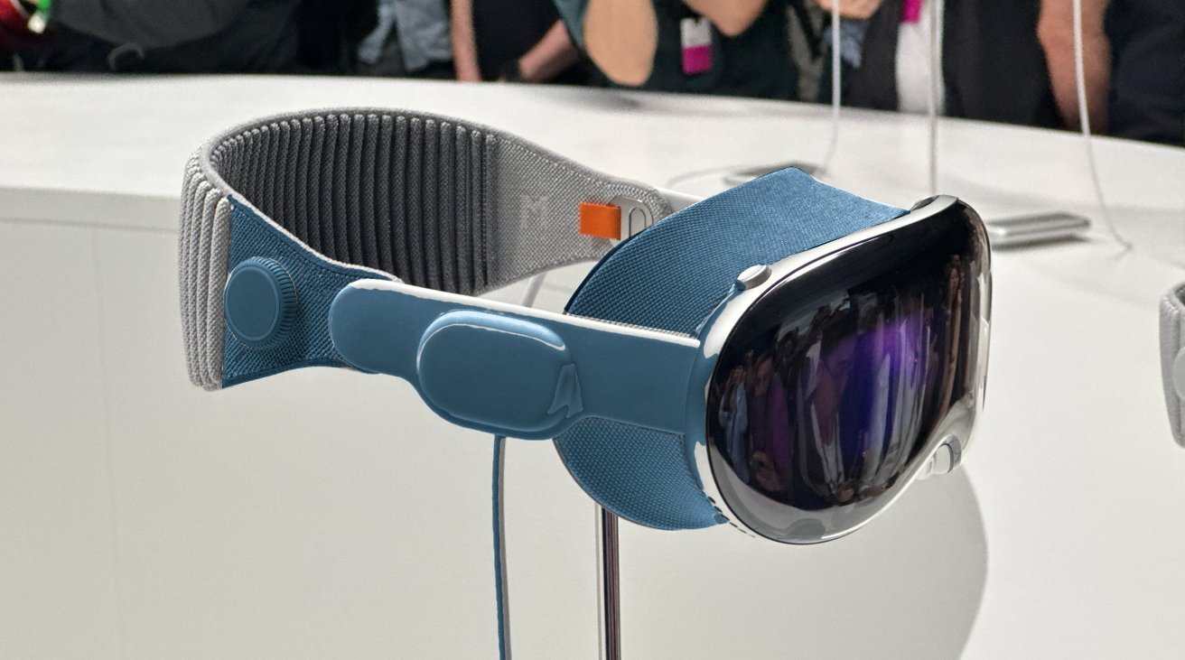 A futuristic headset featuring a reflective visor and adjustable head strap displayed on a stand, with people blurred in the background.