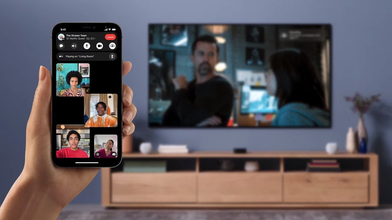 A hand holds a smartphone showing a video call with four people. A blurred TV in the background displays a scene with two characters.