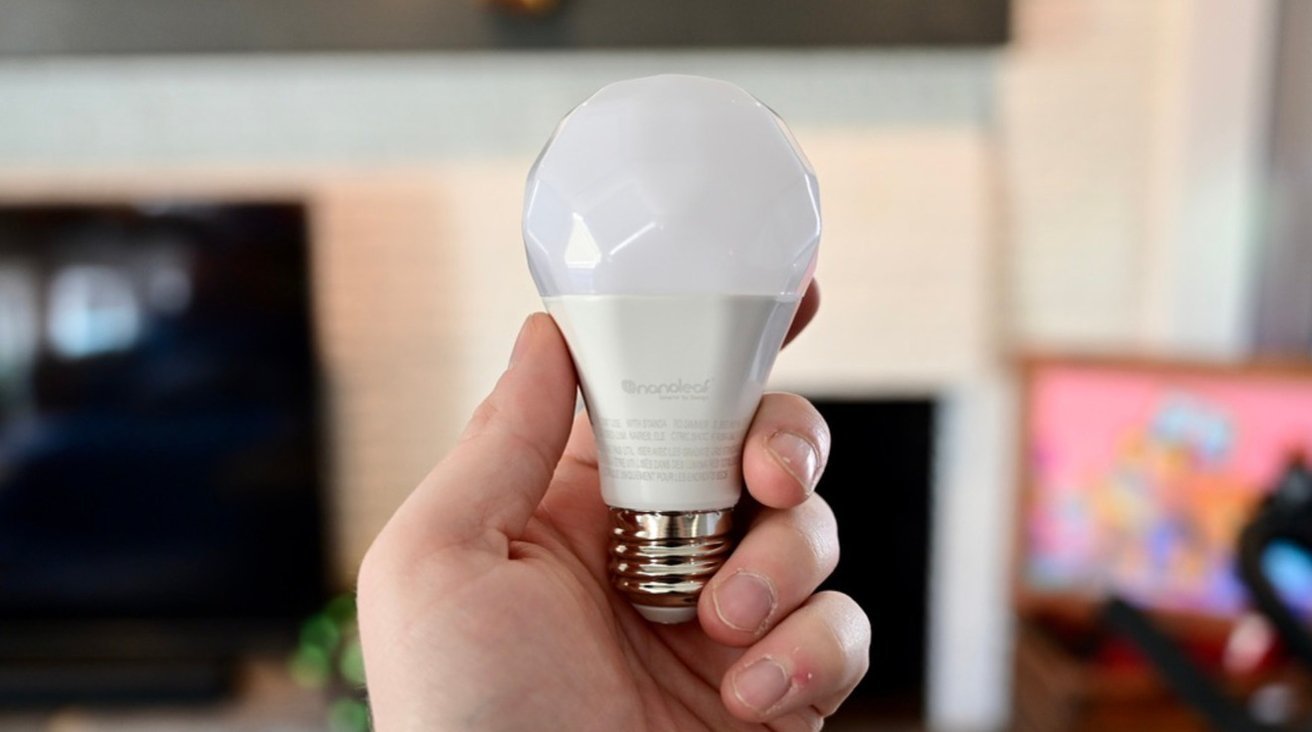Hand holding a faceted white LED light bulb, with a blurred indoor background including a television and colorful artwork.