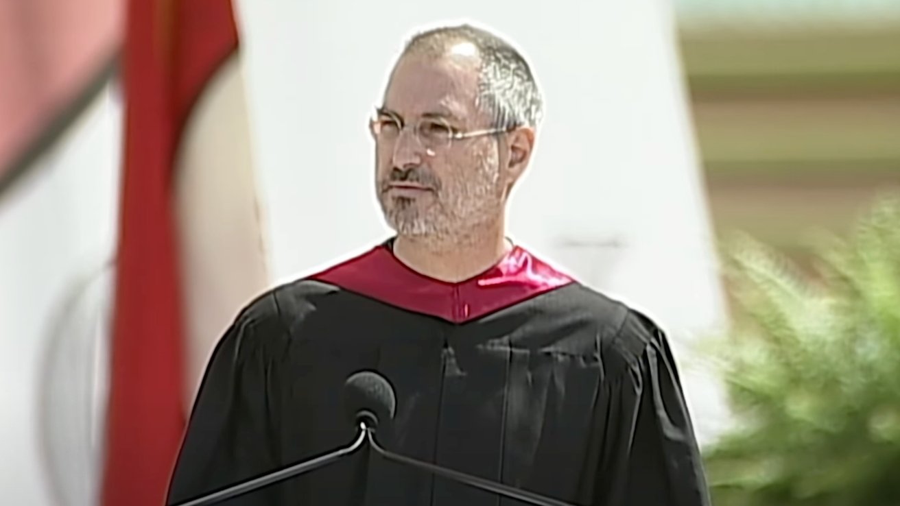 Person in black and red academic gown speaking at an outdoor event, with foliage and a blurred background.