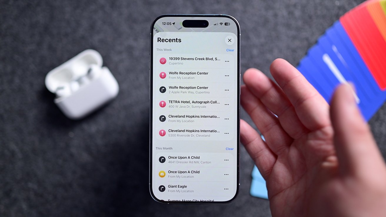 A person holds a smartphone displaying a recent locations list. Nearby are wireless earphones in their open case on a textured surface with colorful cards in the background.
