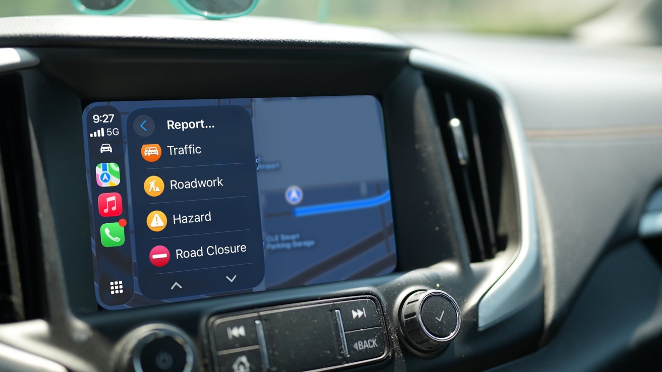 Car dashboard screen displaying navigation with report options for traffic, roadwork, hazards, and road closures. Surrounding interior includes control knobs and buttons.