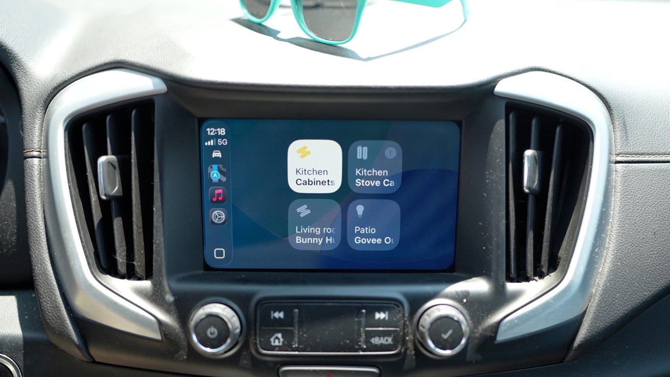 Car dashboard screen displaying home automation controls with options for kitchen cabinets, stove, living room, and patio. Sunglasses rest on top, with sunlight highlighting details.