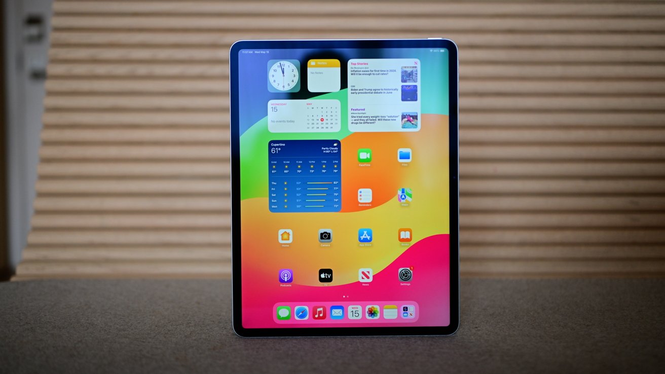 Tablet displaying colorful home screen with app icons, widgets, and clock, resting on a table against a striped background.