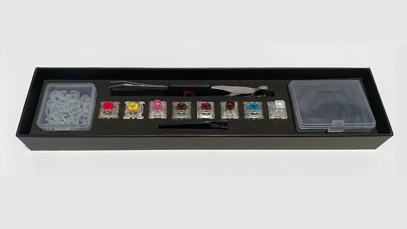 Keyboard switch kit containing various colorful switches, O-rings, tweezers, brush, and cleaning cloth in a black box.