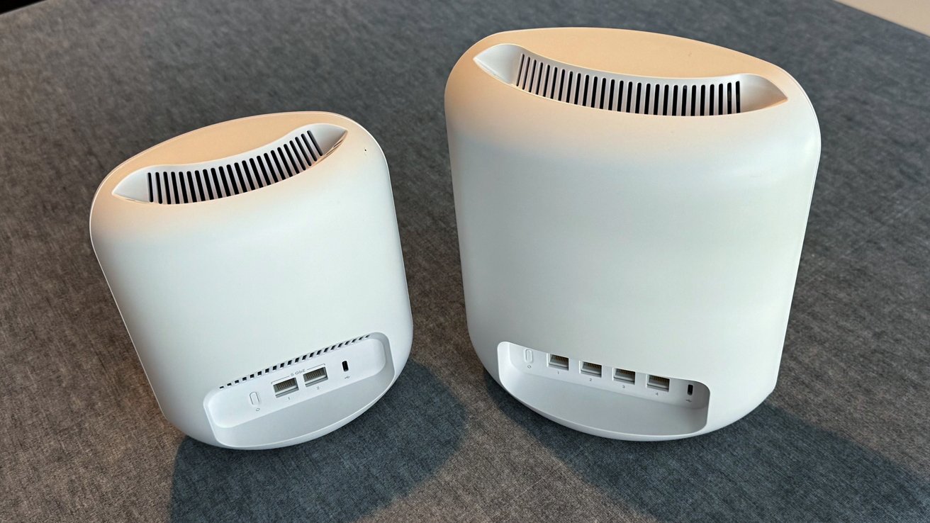 Two white, cylindrical electronic devices with ventilation slits on top and multiple ports on the back, placed side by side on a gray surface.