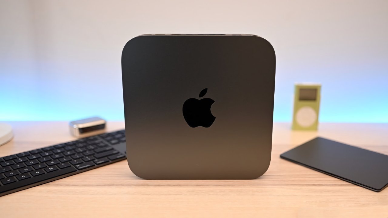 Black compact computer device with Apple logo on a wooden desk, surrounded by a keyboard, small electronic devices, and soft ambient lighting.