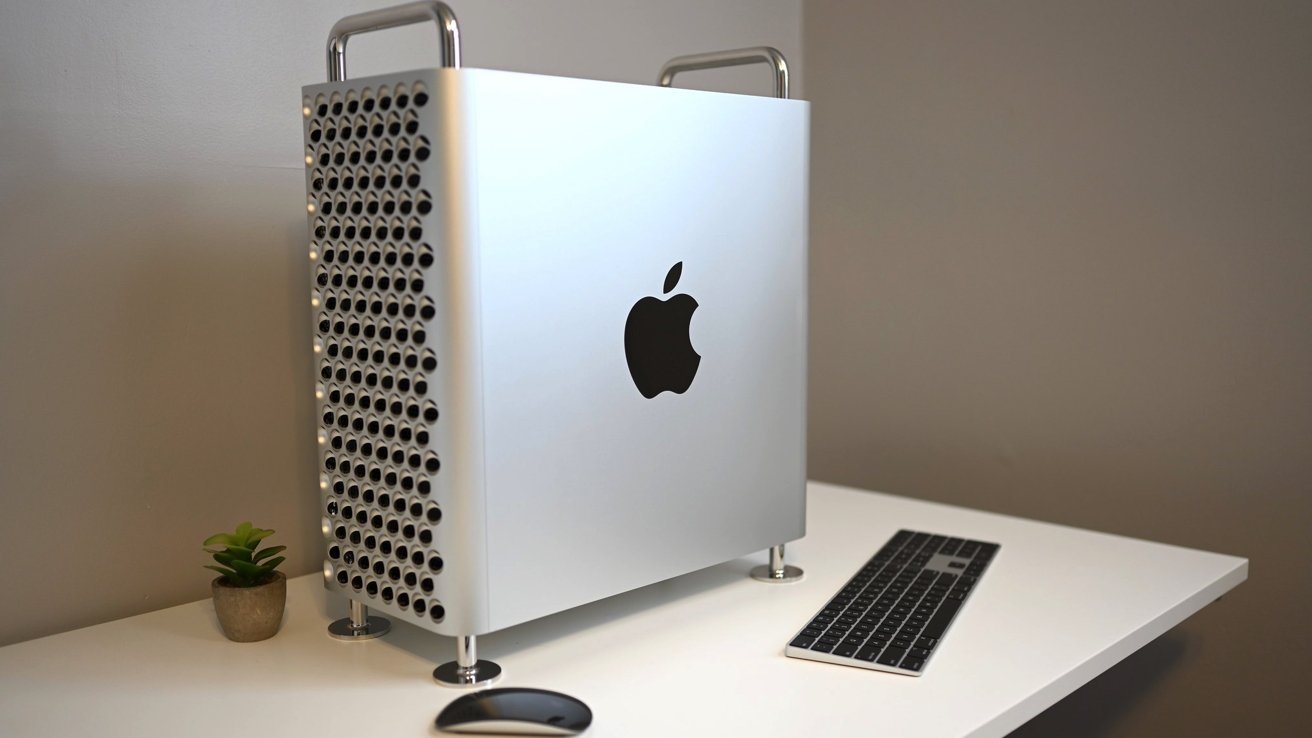 Silver computer tower with a perforated side, black apple logo, silver handles, on a white desk with a small plant, keyboard, and mouse.