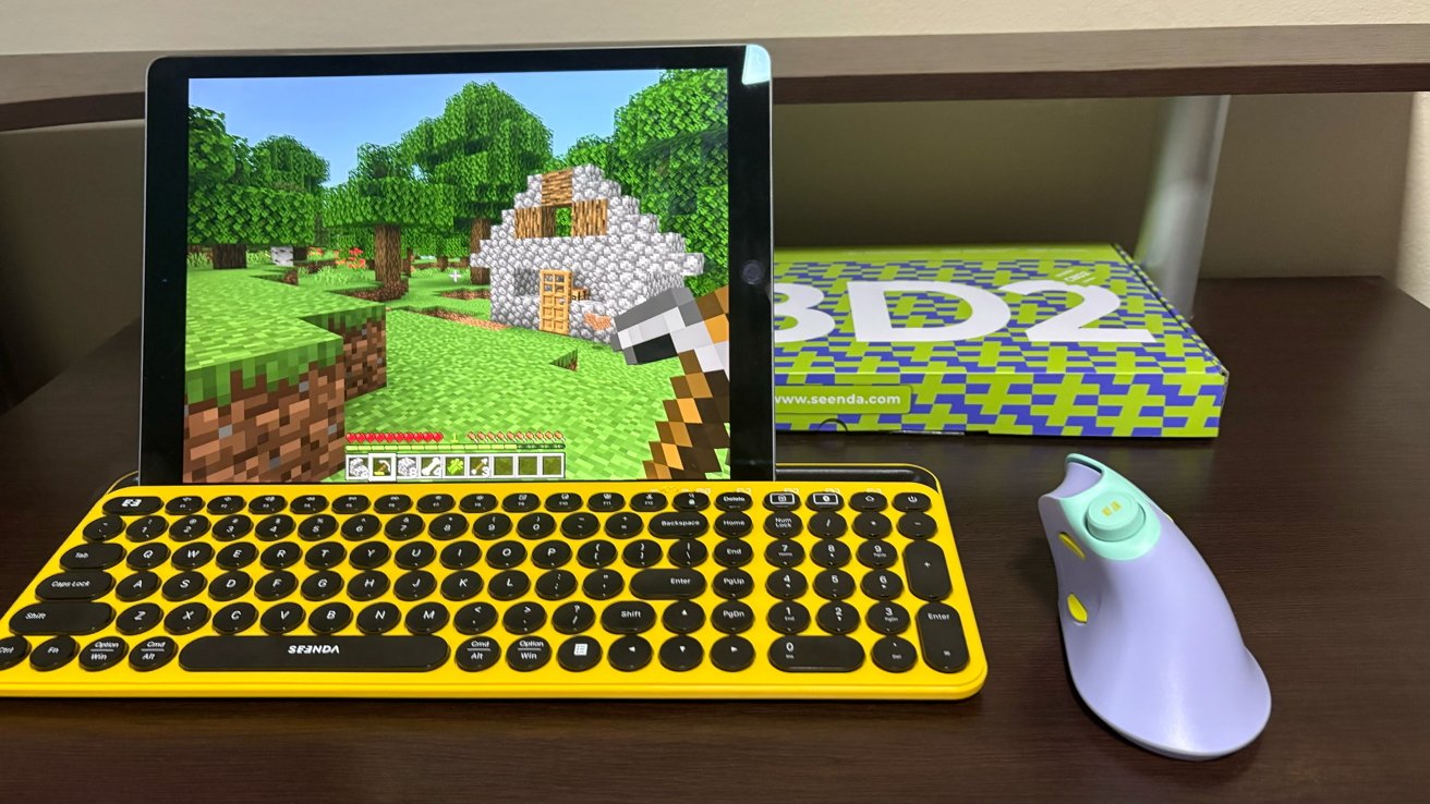 Tablet displaying a block-based video game, yellow keyboard below, and purple mouse beside on a dark brown desk. Box with geometric design and text in the background.