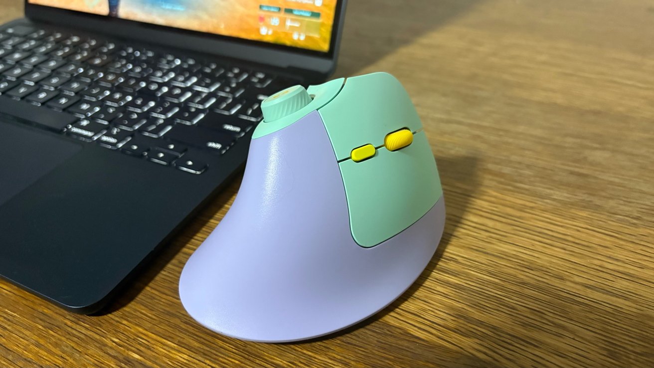 Ergonomic vertical mouse with purple and mint green design, yellow scroll wheel and buttons, beside a black laptop on a wooden surface.