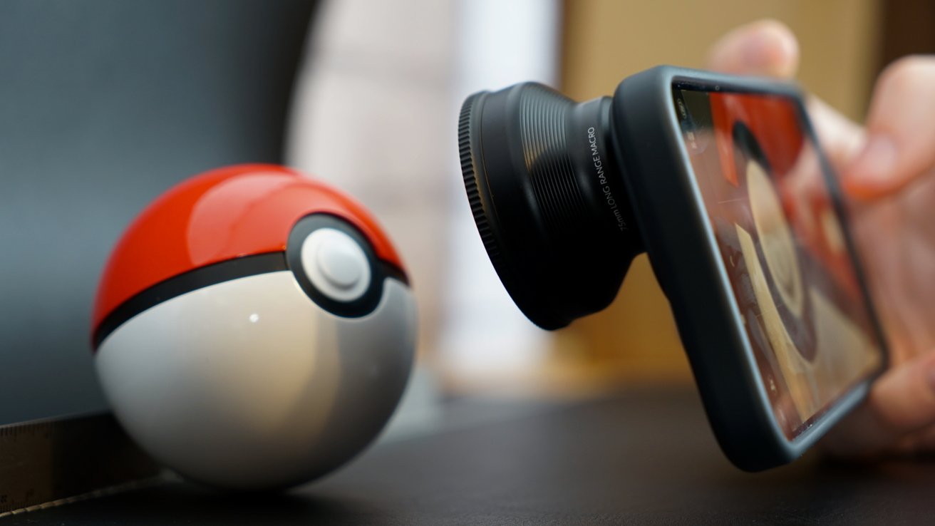 A red and white Poke Ball beside a smartphone with an external lens attachment, capturing a close-up view.