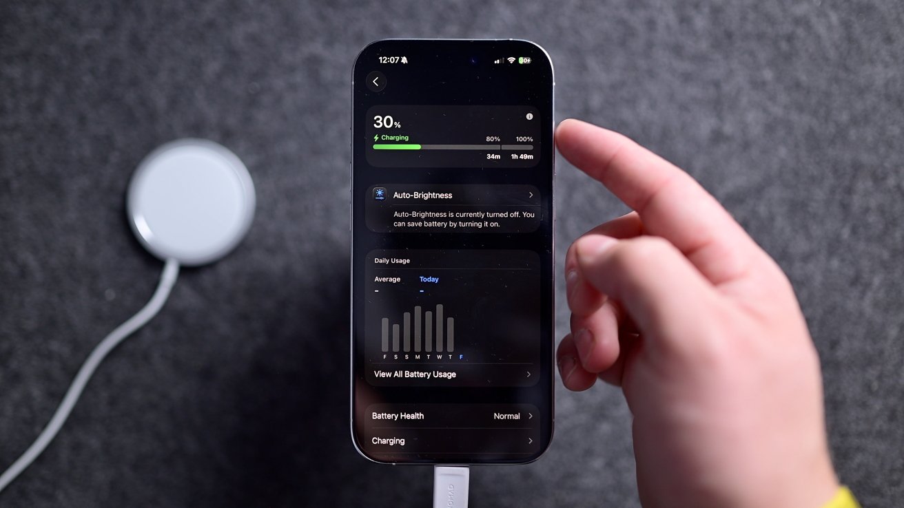 Hand holding a smartphone displaying battery charging status at 30%, alongside a wireless charging pad on a dark textured surface.
