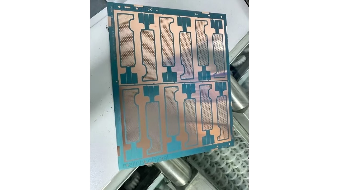 Rectangular circuit board with intricate copper pathways and blue borders, featuring multiple repeating patterns, placed on a white surface near cylindrical components.
