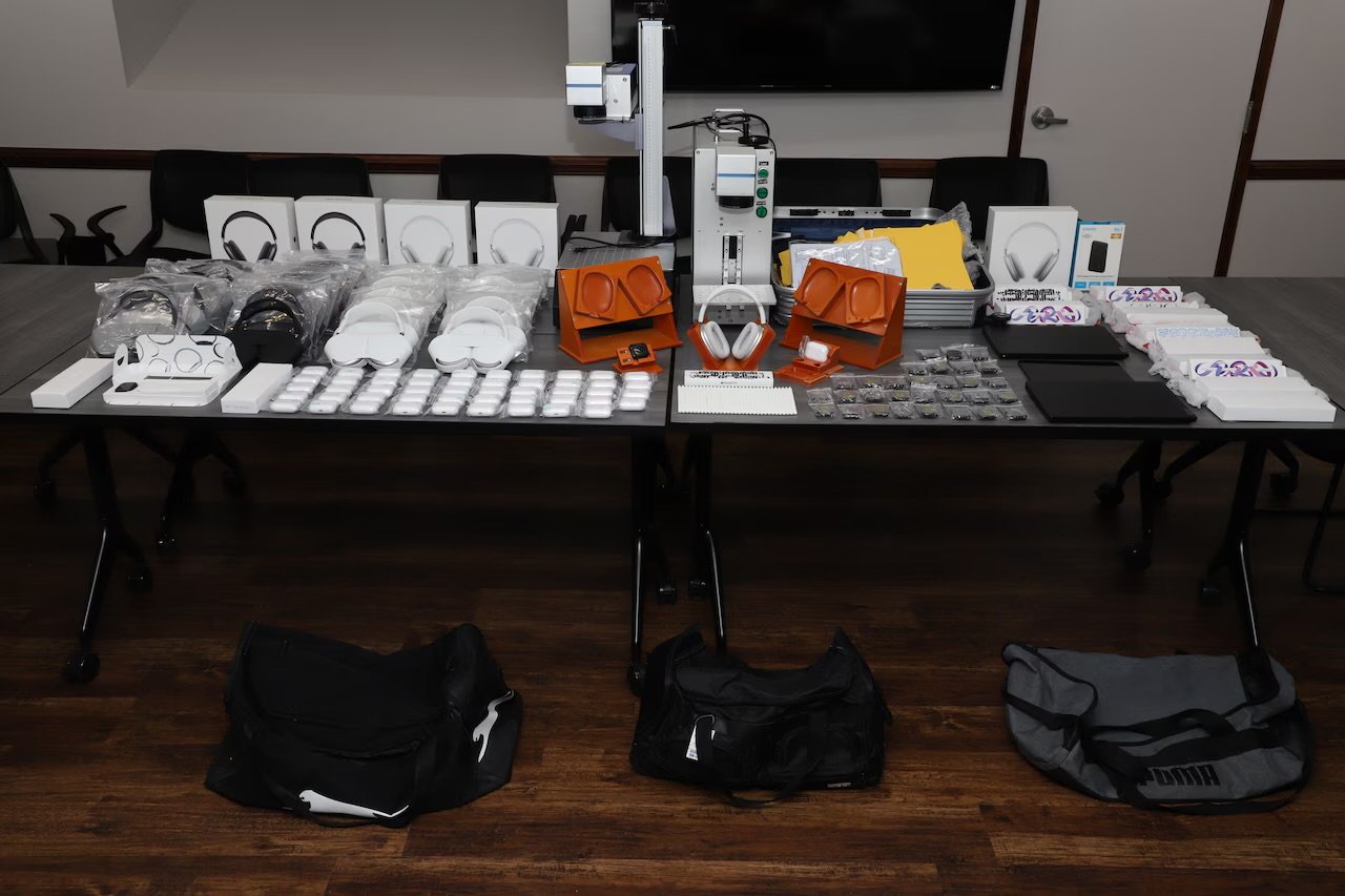 A table with headphones, packaged electronics, orange stands, bags, and a machine, arranged neatly in a room with wooden flooring and dark chairs.