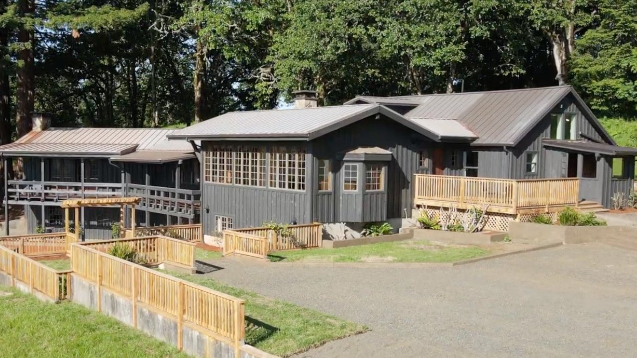 A rustic, dark gray wooden house with large windows, surrounded by a wooden fence and lush greenery, under a clear blue sky.