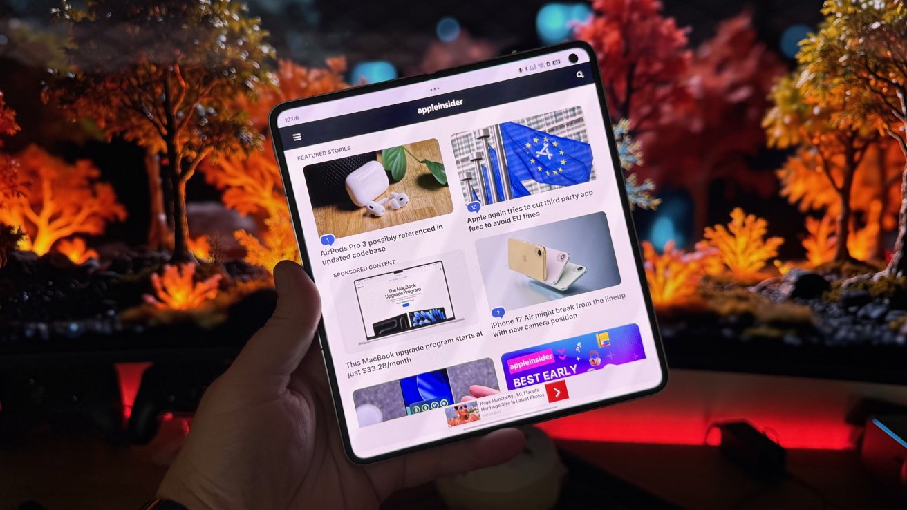 A hand holds a touchscreen device displaying a tech news website with various articles, set against a vibrant, blurred background of trees with orange and red leaves.