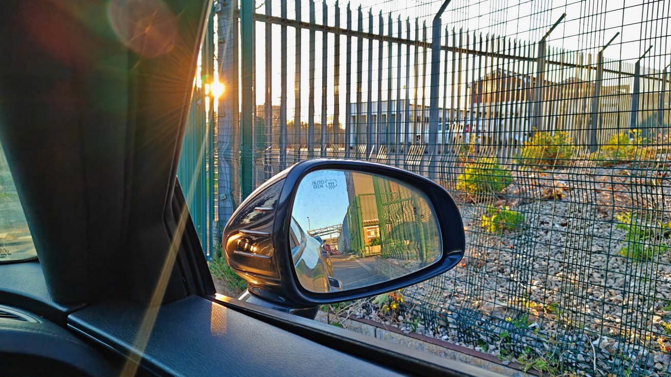 Car side mirror reflecting a street scene; foreground shows sunlight peeking through a metal fence, with some greenery and rocks.