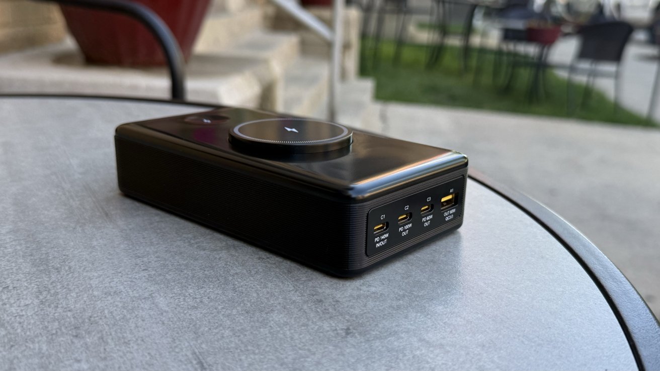 Portable black charging device with multiple ports, placed on a round metal table outdoors. Steps and chairs are visible in the background.